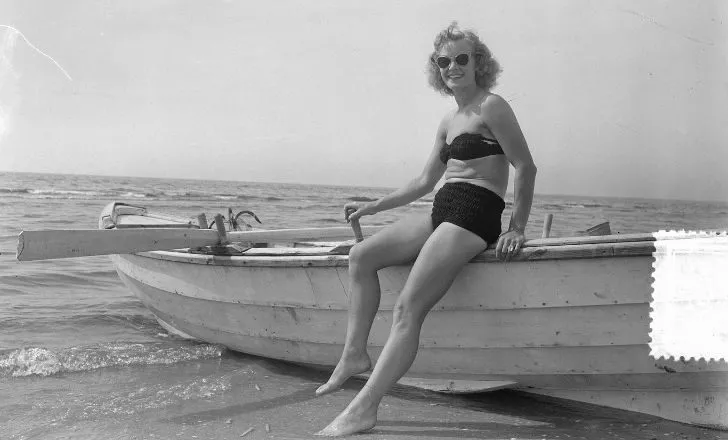 A woman sitting on a boat wearing a bikini on the beach in the 1930's.